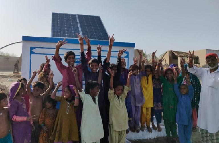 Happy kids stood in front of a solar-powered well as part of a water project